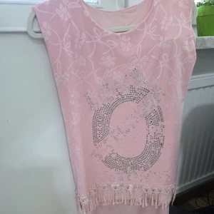 Rosa kortklänning med paljetter och fransar - Söt rosa kortklänning med blommigt mönster i tyget, glittrande paljettdetaljer framtill och dekorativa fransar längst ner. Ärmlös modell med rund halsringning, perfekt för dig som gillar en feminin och lekfull stil.