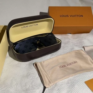 Svarta Louis Vuitton solglasögon - Snygga svarta solglasögon från Louis Vuitton med breda skalmar och LV-monogram på glasen. Glasögonen har en modern, rektangulär form och levereras med originalfodral och putsduk. Perfekta för dig som vill sticka ut med en lyxig accessoar.