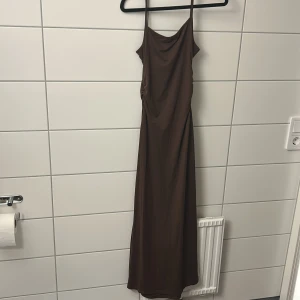 Brun långklänning med tunna axelband - Säljer en stilren brun långklänning med tunna axelband och snygg drapering vid bysten. Klänningen har en smickrande passform och faller snyggt längs kroppen. Perfekt för dig som gillar minimalistisk och trendig stil.