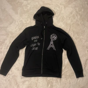 Svart rhinestone hoodie från blinglucid  - Svart hoodie från BLINGLUCID med huva och dragkedja. Framsidan har strasstenar som bildar Eiffeltornet och texten 'paris au clair de lune'. Mjuk insida och långa ärmar, perfekt för dig som gillar streetstyle med lite extra bling.
