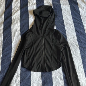 Svart croppad hoodiejacka med dragkedja - Svart croppad hoodiejacka med dragkedja framtill och långa ärmar. Jackan har en figurnära passform och är tillverkad i ett ribbat syntetmaterial. Perfekt för en sportig och trendig look.