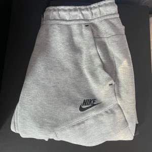 Grå mjukisbyxor från Nike - Gråa mjukisbyxor från Nike med svart logga på benet. Byxorna har elastisk midja med svarta snören och praktiska fickor. Tillverkade i mjukt bomullsmaterial och har en svart detalj på ena benet. Perfekta för chill eller träning.
