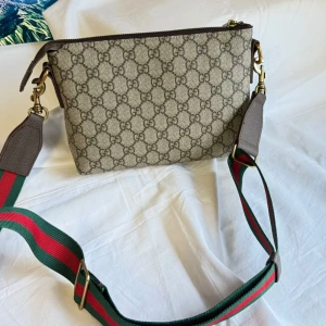 Gucci axelväska med GG-mönster - Snygg axelväska från Gucci i beige och brunt GG-mönster. Väskan har bred, justerbar axelrem i grönt och rött, klassiska Gucci-färger. Tillverkad i canvas med detaljer i brunt skinn och dragkedja upptill. Perfekt storlek för det viktigaste och med innerfack för småsaker.