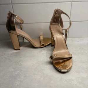 Guldiga sandaler med klack och öppen tå - Snygga sandaler i metallic guld med bred klack och öppen tå. Rem runt vristen med spänne och stilren design. Perfekta för dig som vill sticka ut med ett par glammiga skor. Skorna har en plattformsula framtill och är tillverkade i syntetmaterial.