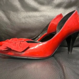 Klarröda 50-talsklackar i lack med mockarosett - stl 35 - Pinup - Retro - Rockabilly  -  Äkta vintagepumps i klarrött lackläder med söt rosett i mocka framtill. Spetsig tå och smal klack (ca 7 cm) för en klassisk siluett. Perfekta för dig som gillar pinup, rockabilly eller retrostil. Storlek 35, innerlängd ca 23 cm. Mycket fint skick. Jag prutar inte – priset är redan lågt – men erbjuder rabatt vid köp av flera saker. Rensar garderoben – kika gärna på mina andra annonser också!