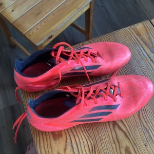 Röda Adidas fotbollsskor med dobbar - Säljer ett par röda Adidas F50 fotbollsskor med svarta detaljer och klassiska tre ränder på sidorna. Skorna har snörning och dobbar för bra grepp på planen. Ovandelen är i syntetmaterial och insidan är svart. Perfekta för dig som vill ha en par sköna och hyfsat lätta skor, Billigare vid snabb affär