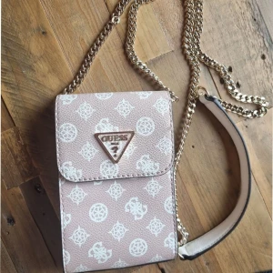 Guess mobilväska  - Guess Noelle Girlfriend mobilväska i färgen Rosewood med guldfärgad kedjan.  Mått: ca 11 x 17 x 3,5 cm Smartphone-fack: ca 9 x 16 x 3,5 cm Axelrem: ca 123 cm Material: PU på utsidan, polyester/bomull  Egenskaper  Stängs med klaff och magnetlås 3 kortfack i huvudfacket 1 extra insticksfack på baksidan  OBS! Väskan är använd flitigt, därför finns mindre defekter/slitage (se bilder), men den har varit väl omhändertagen och är i fullt användbart skick. Nypris: ca 1 300 kr.