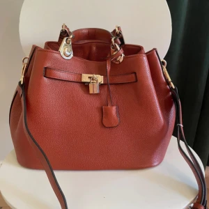 Brun handväska från Noble Bags - Snygg brun handväska från Noble Bags med guldfärgade metalldetaljer och lås framtill. Väskan har både handtag och axelrem, vilket gör den enkel att bära. Stilren design med struktur i materialet och praktiska fack inuti. Jag har gjort extra hål i axelremmen för att kunna göra den kortare. Den är 35 cm bred, 20 cm djup och 24 cm hög.