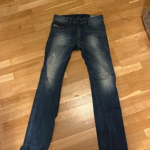 Blå slitna jeans från Diesel med raka ben - Säljer ett par blå jeans med snyggt slitna detaljer och raka ben. Jeansen har klassisk femficksdesign, normal midja och ljusare partier på låren och knäna för en cool, använd look. Perfekta för dig som gillar en avslappnad stil.