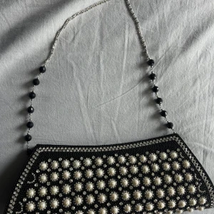 Svart kuvertväska med pärlor och kedja - En svart kuvertväska dekorerad med vita pärlor och glittrande stenar på framsidan. Väskan har en silverfärgad kedja med svarta pärldetaljer som axelrem. Stängs med dragkedja och har ett stilrent, avlångt format. Perfekt för att lyfta din outfit med extra bling.