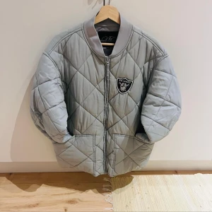 Quiltad bomberjacka Urban Classics XL - Snygg ljusgrå quiltad bomberjacka från Urban Classics i storlek XL. Jackan har Raiders-logga på bröstet, dragkedja framtill och två stora fickor. Perfekt för dig som gillar sportig och street-inspirerad stil.