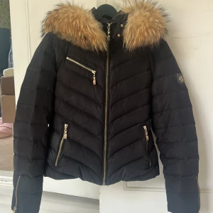 Svart pufferjacka med fuskpäls från Hollies - Snygg svart pufferjacka från Hollies med guldig dragkedja och detaljer. Jackan har en stor huva med fluffig fuskpäls, tre dragkedjefickor framtill och emblem på ärmen. Perfekt för kalla dagar när du vill vara både varm och stilren. Jackan har en liten fläck framtill som tyvärr inte går bort