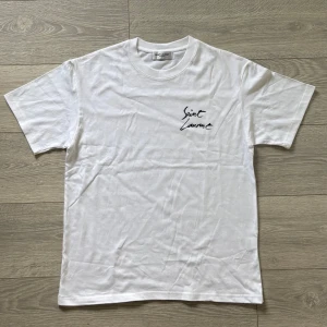Vit Saint Laurent t-shirt med tryck - Vit t-shirt från Saint Laurent med svart grafiskt tryck på bröstet och ryggen. Klassisk rund hals och korta ärmar. Materialet är mjuk bomull som känns skönt mot huden. Perfekt för dig som gillar stilrena designer med en lyxig touch.