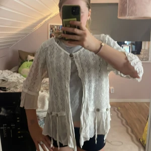 Vit spetskofta från Ambra - Säljer en vit kofta från Ambra med snygga spetsdetaljer och vågigt mönster. Koftan har trekvartsärm, knappar framtill och dekorativa snören vid nederkanten. Lätt och luftig, perfekt för vår och sommar. Made in Italy.