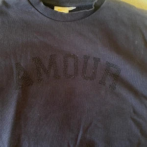 Mörkblå sweatshirt från Gina Tricot - Säljer en mörkblå sweatshirt från Gina Tricot med texten 'AMOUR' i svarta pärlor på bröstet. Tröjan har rund hals, ribbade muddar och är långärmad. Perfekt för dig som gillar stilrena detaljer och vill ha en chill look.