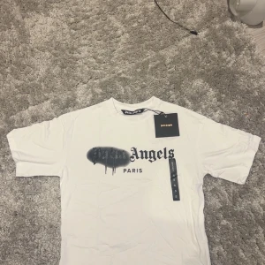 Vit Palm Angels t-shirt Paris print - Vit t-shirt från Palm Angels med Paris-tryck på bröstet och coolt svart spray-effekt över loggan. Klassisk rund hals och korta ärmar. Materialet är mjuk bomull och passformen är normal. Perfekt för dig som gillar streetwear och snygga detaljer.