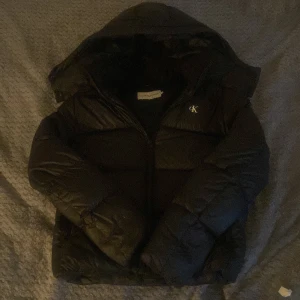 Svart pufferjacka Calvin Klein Jeans - Svart pufferjacka från Calvin Klein Jeans med huva och dragkedja. Jackan har diskret CK-logga på bröstet och är vadderad för extra värme. Perfekt för kalla dagar och har en clean, modern look som funkar till allt. Om du har några funderingar eller vill ha bättre bilder kom privat, och priset kan diskuteras vid snabba affärer 