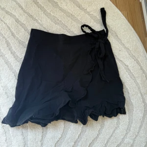 Svart kjol - Svart kortkjol med omlott och knyt i midjan. Kjolen har volangkant längst ner. Är helt oanvänd