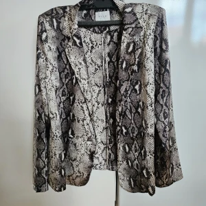 Ormskinnsmönstrad kavaj från Vila - Säljer en snygg kavaj från Vila med ormskinnsmönster i svart, grått och beige. Kavajen har klassisk krage, lång ärm och en loose fit. Perfekt för dig som vill sticka ut med en trendig look.