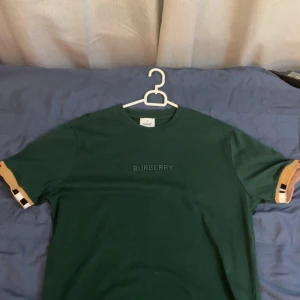 Grön Burberry t-shirt med logga - Mörkgrön t-shirt från Burberry med diskret logga på bröstet och klassiska Burberry-rutiga detaljer på ärmsluten. T-shirten har rund hals och är kortärmad, perfekt för en clean och stilren look.