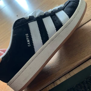 Adidas Campus svart/vit storlek 40 - Snygga Adidas Campus sneakers i svart mocka med klassiska vita ränder och vit sula. Du får med svarta skosnören också. Köpte skorna för 1000kr och dom är använda 1 gång.