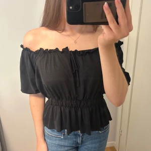 Svart offshoulder blus med volang - Säljer en svart offshoulder blus med korta puffärmar och volangkant nertill. Toppen har resår i midjan och runt axlarna samt en söt knytning framtill. Använd fåtal gånger💕