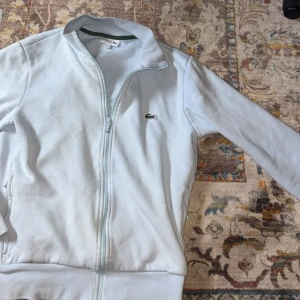 Ljusblå zipjacka från Lacoste - Ljusblå zipjacka från Lacoste med klassisk krage och dragkedja framtill. Jackan har lång ärm och den ikoniska krokodilloggan broderad på bröstet. Perfekt för en chill och sportig stil.