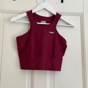 Vinröd linnetopp från Stronger - Snygg vinröd linnetopp från Stronger med sportig look och hög halsringning. Toppen är ärmlös och tillverkad i ett stretchigt syntetmaterial som passar perfekt till träning. Diskret logga framtill och bred resår nedtill för extra stöd.