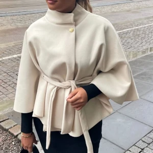 Beige kappa med bälte och vida ärmar - Stilren beige kappa med hög krage, knäppning upptill och knytskärp i midjan. Kappan har vida ärmar och en elegant, minimalistisk look. Perfekt för dig som gillar en clean och sofistikerad stil🥰orginal pris 538 ( ink frakt ). Använd 1 gång. Kappan har en liten fläck på ryggen, men kom så när jag beställde den☺️