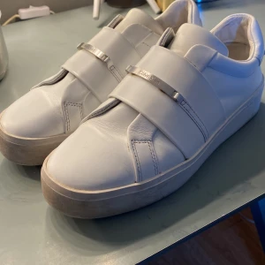 Vita sneakers från Calvin Klein - Stilrena vita sneakers från Calvin Klein i skinn med breda remmar och metallspännen med logga. Snygg minimalistisk design och rund tå, perfekt för dig som gillar cleana och moderna skor. Notera att skorna har en hög sula💕