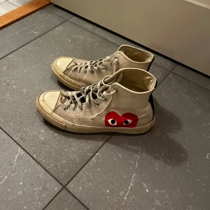 Converse x CDG Play vita high sneakers - Säljer ett par Converse Chuck Taylor All Star high sneakers i samarbete med Comme des Garçons Play. Skorna är vita i canvas med den ikoniska röda hjärtloggan på sidan, svarta detaljer baktill och klassisk snörning. Perfekta för dig som gillar streetstyle och vill sticka ut.