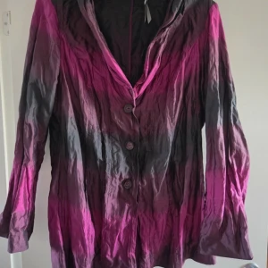 Svart och rosa kavaj i metallic - Unik kavaj i svart och rosa med metallicskimmer och snygg skrynklig effekt. Långa ärmar, tre knappar framtill och coolt färgskiftande tyg. Perfekt för dig som vill sticka ut med en statement-piece. Materialet är polyester och metallfibrer.