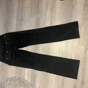 Juicy couture byxor - Säljer ett par svarta mjukisbyxor i mjuk sammet med breda ben och dragsko i midjan. Byxorna har fickor med lock och knappar bak, vilket ger en chill och stilren vibe. Perfekta för dig som gillar bekväma och snygga byxor med lite extra attityd.