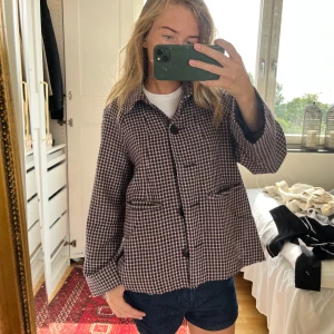 Rutig overshirt med knappar - Snygg rutig overshirt i mörkbruna och beige toner med klassisk krage och stora svarta knappar framtill. Jackan har två fickor fram och en dekorativ knappdetalj bak. Passformen är loose och materialet känns som bomull eller ullblandning.