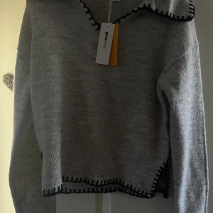 Grå stickad tröja från Gina Tricot - Mysig grå stickad tröja från Gina Tricot med svarta dekorativa sömmar längs v-ringningen och nederkanten. Tröjan har lång ärm och ribbade muddar, perfekt för lager på lager under kyliga dagar. Kvar prislappen så aldrig använd. köpt på gina young Storlek 170 funkar som Xs