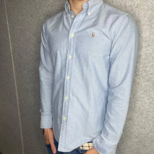 Ljusblå skjorta från Ralph Lauren - Klassisk ljusblå skjorta från Ralph Lauren med broderad logga på bröstet. Skjortan har button-down krage, långa ärmar och stängs med vita knappar framtill. Tillverkad i mjuk bomull som känns skön mot huden. Perfekt för en clean och stilren look.