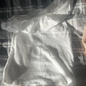 Vit oneshoulder topp med volang - Superfin vit topp med oneshoulder-design och bred volang längs halsringningen. Tunt, ribbat tyg som ger en snygg struktur. Perfekt för dig som vill sticka ut med en trendig och stilren look.