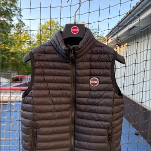 Svart dunväst från Colmar - Snygg svart dunväst från Colmar med quiltad design och hög krage. Västen har dragkedja framtill, två fickor med dragkedja och Colmar-logga på bröstet. Perfekt att slänga över en hoodie för en chill look.