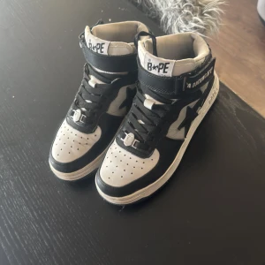 Svarta och vita Bape Sta sneakers - Säljer ett par klassiska Bape Sta sneakers i svart och vitt skinn. Modellen är high-top med snörning, perforerad tå och ikonisk stjärndetalj på sidan. Skorna har BAPE-logga på plösen och kardborreband vid ankeln för extra streetkänsla.