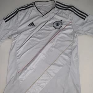 Tysklands vit Adidas fotbollströja M - Vit officiell Tyskland fotbollströja från Adidas med svarta, röda och guld diagonala linjer på framsidan. Tröjan har svarta Adidas-ränder på axlarna, broderat landslagsmärke och ClimaCool-material som andas. Snygg V-ringning och tryck i nacken med 'Deutscher Fußball-Bund'.
