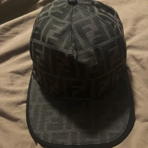 Svart Fendi keps med logomönster - Snygg svart keps från Fendi med det klassiska FF-logomönstret över hela kepsen. Kepsen har böjd skärm och justerbar rem baktill. Tillverkad i ett slitstarkt tyg med exklusiv känsla och diskreta detaljer. Perfekt för dig som vill sticka ut med en lyxig accessoar. Strapen är lös men om du har storlek M så är det inga problem 