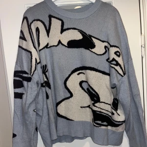 Ljusblå oversized stickad tröja med tryck - Säljer en ljusblå oversized stickad tröja med stort grafiskt tryck i svart och vitt. Tröjan har rund halsringning och långa ärmar. Perfekt för dig som gillar streetstyle och vill sticka ut med en cool design.