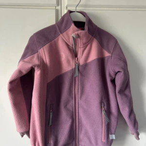 Lila och rosa fleecejacka från PAX - Fleecejacka från PAX i lila och rosa med dragkedja framtill och två fickor med dragkedjor. Jackan har hög krage och är perfekt för kyliga dagar. 
