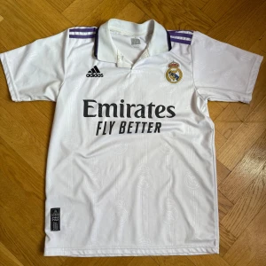 Real Madrid Benzema matchtröja Adidas - Snygg vit Real Madrid fotbollströja från Adidas med Benzema och nummer 9 på ryggen. Klassisk krage med lila detaljer på axlarna och broderad klubbmärke på bröstet. Tillverkad i lätt och ventilerande material, perfekt för fotbollsplanen.