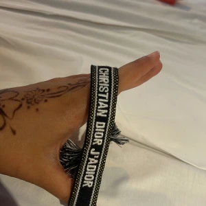 Christian Dior svart tygarmband - Svart och vitt armband från Christian Dior med texten 'CHRISTIAN DIOR J'ADIOR' broderad. Armbandet är gjort i tyg med flätade band och tofsar i ändarna. Snygg accessoar med exklusiv känsla och tydlig logga.