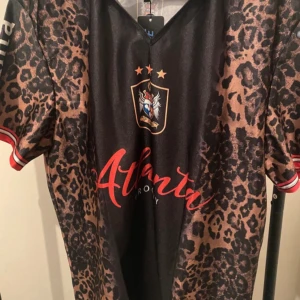 Atlanta Groovy leopard tröja  XL - Unik tröja från ROMWE i storlek XL med svart bas och leopardmönstrade ärmar. Tryck med Atlanta Groovy och klubbmärke på bröstet. Röd text och detaljer på ärmsluten. Tillverkad i lätt och glansigt material, perfekt för dig som vill sticka ut.