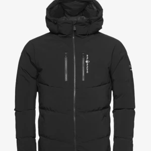 Svart dunjacka från Sail Racing - Svart lång dunjacka från Sail Racing med huva och dragkedja framtill. Jackan har en clean look med diskreta loggor på bröstet och ärmen, samt praktiska fickor. Perfekt för kalla dagar när du vill hålla stilen och värmen.