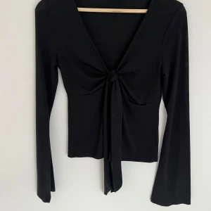 Svart omlottopp med knyt från Gina Tricot - Svart långärmad omlottopp från Gina Tricot i storlek S. Toppen har en djup v-ringning och snygg knytning framtill. Materialet är mjukt och stretchigt, perfekt för att styla med jeans eller kjol.