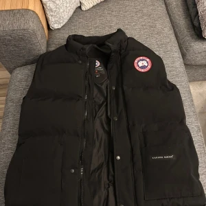 Svart dunväst från Canada Goose - Säljer en svart dunväst från Canada Goose med klassisk logotyp på bröstet och diskret ficka framtill med varumärkesnamn. Västen har hög krage och stängs med både dragkedja och knappar. Perfekt för kyliga dagar. Liten defekt då två knappar lossnat men inget man tänker på! Vid frågor eller funderingar är det bara att höra av sig!