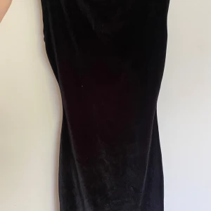 Svart långärmad sammetsklänning - Snygg svart långklänning i mjuk sammet med djup v-ringning både fram och bak. Klänningen är ärmlös och har en elegant, figurnära passform som ger en stilren siluett. Perfekt för dig som gillar minimalistisk och klassisk stil.